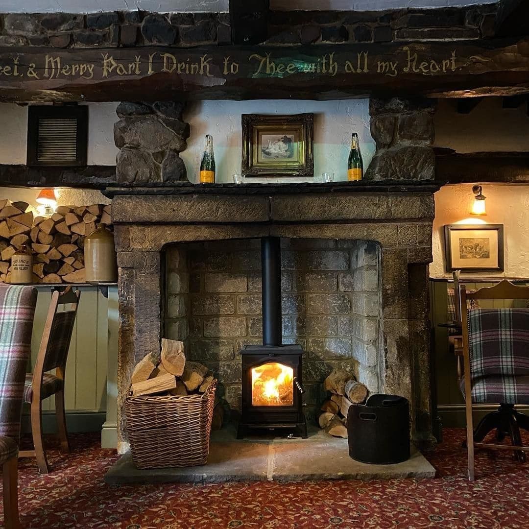 Cozy fireplace at Scotts Arms with a lit wood stove, stone surround, and a basket of logs, flanked by traditional chairs and vintage decor.