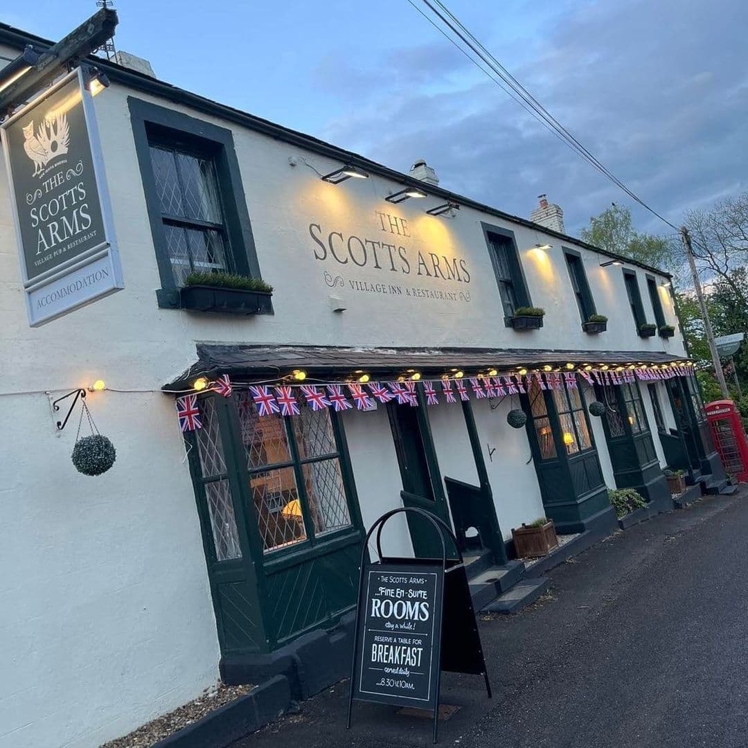 Exterior view of The Scotts Arms, a traditional village inn and restaurant with a white facade, adorned with British flags, and a sign advertising rooms and breakfast.