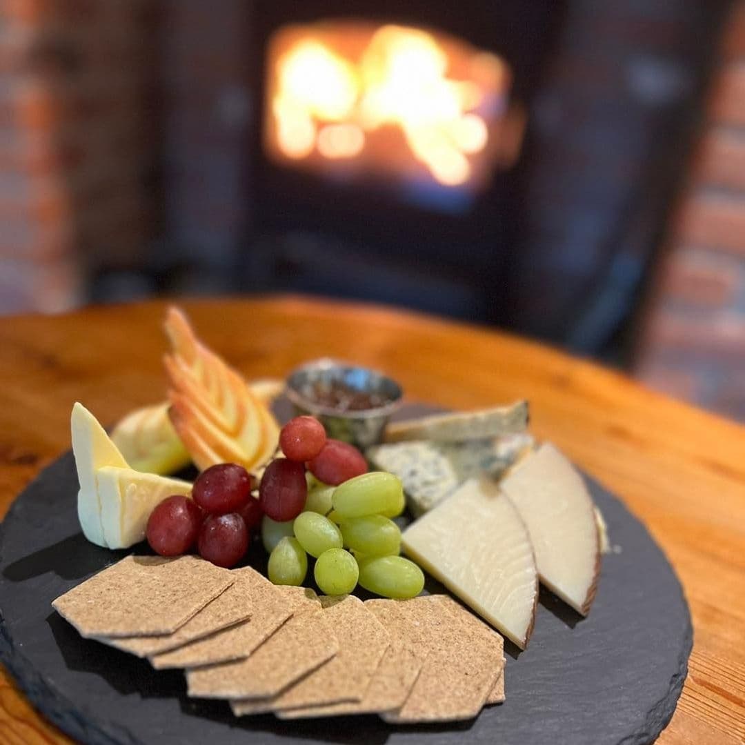 Cheese platter with assorted cheeses, grapes, crackers, and apple slices on a slate board at Scotts Arms, with a cozy fireplace in the background.