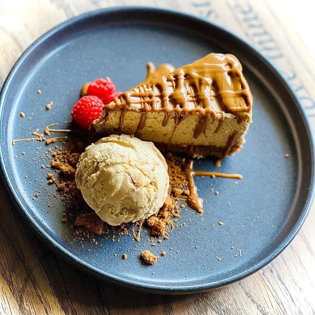 Decadent caramel-drizzled cheesecake with a scoop of vanilla ice cream and fresh raspberries on a blue plate at The Chequers Inn.