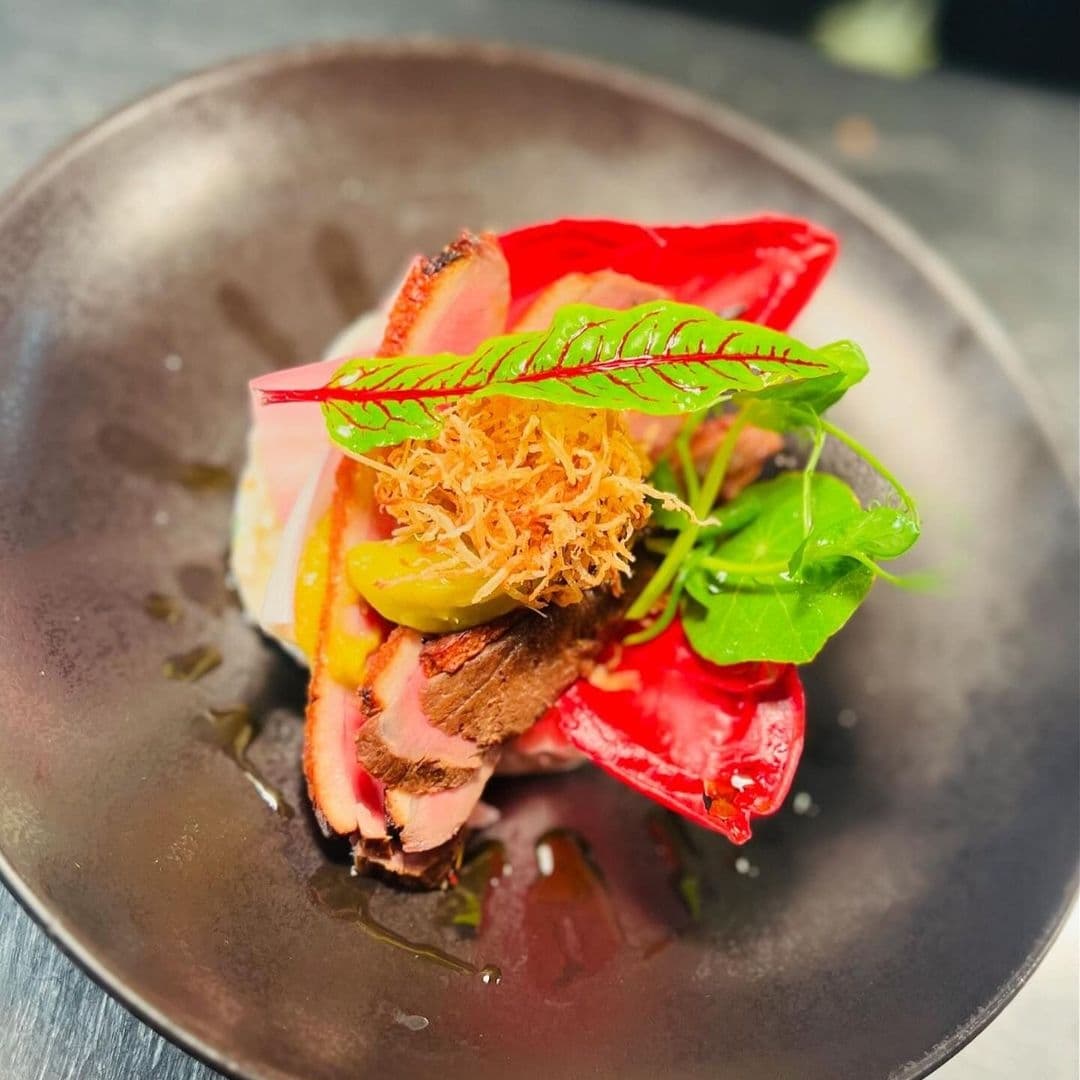 Gourmet dish featuring sliced duck, colorful heirloom tomatoes, green leaves, and crispy fried topping served on a dark plate at The Chequers Inn.