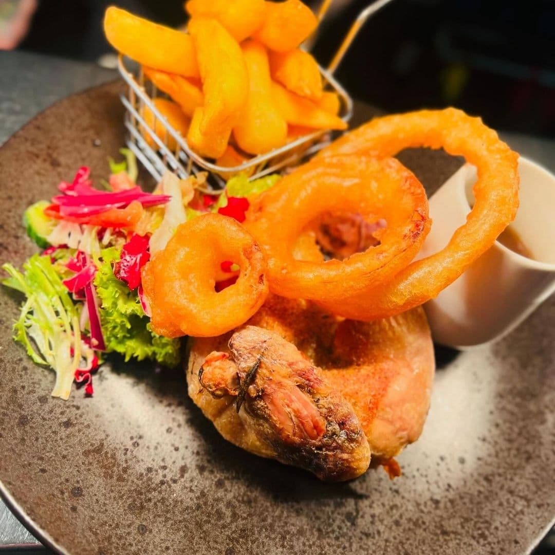 Gourmet meal at The Chequers Inn featuring crispy golden onion rings, a portion of thick-cut chips in a metal basket, a succulent roasted chicken leg, and a side of fresh mixed salad, all served on a stylish ceramic plate.