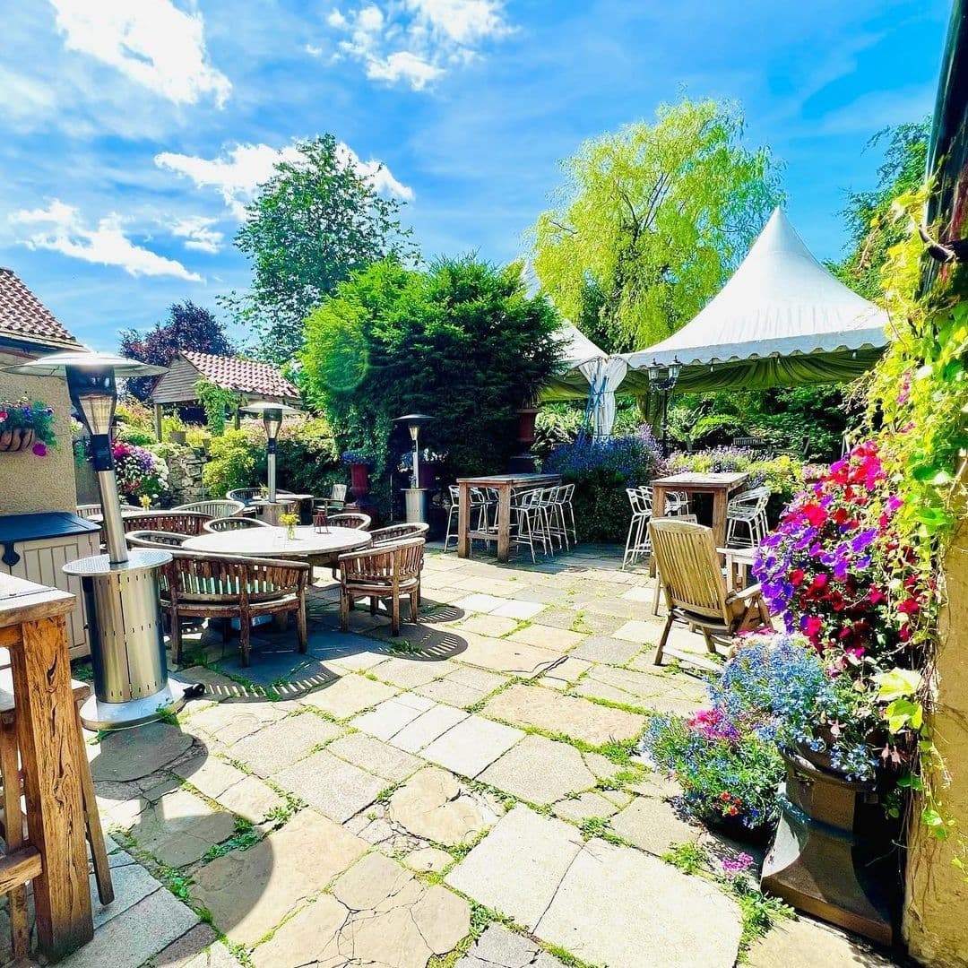Sunny outdoor seating area at The Chequers Inn with wooden tables, chairs, colorful flower arrangements, patio heaters, and a white canopy.