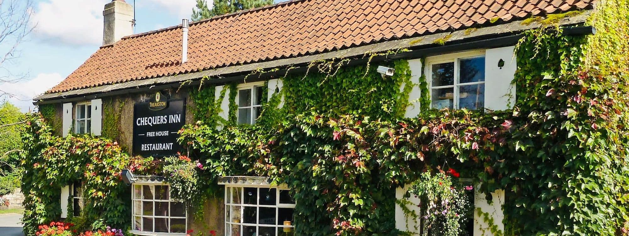 Exterior view of The Chequers Inn, showing a quaint building with a tiled roof, partially covered in green ivy, with a signboard and colorful flowers in front.