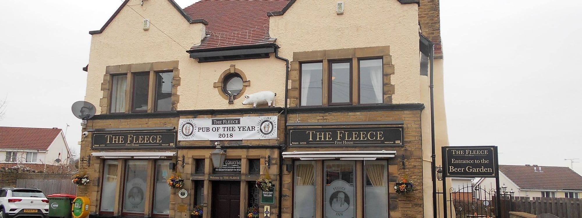 Exterior view of The Fleece, a two-story beige building with a red roof, featuring a sign indicating "Pub of the Year 2018" and a white sheep statue on the roof. The entrance displays the pub's name with hanging flower baskets and a sign pointing to the beer garden.