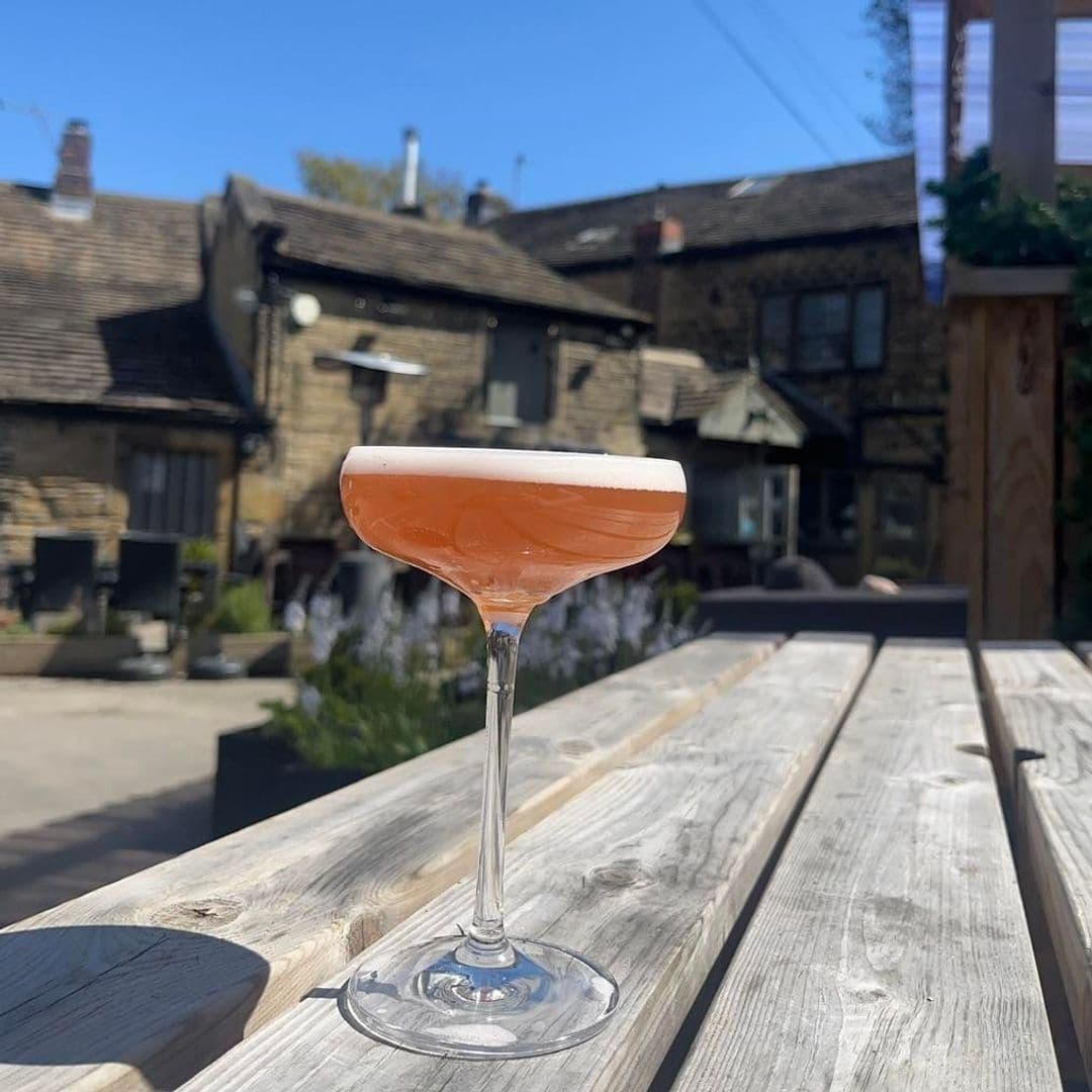 Cocktail on outdoor wooden table at The Thornhill with blurred background of traditional stone building and patio.
