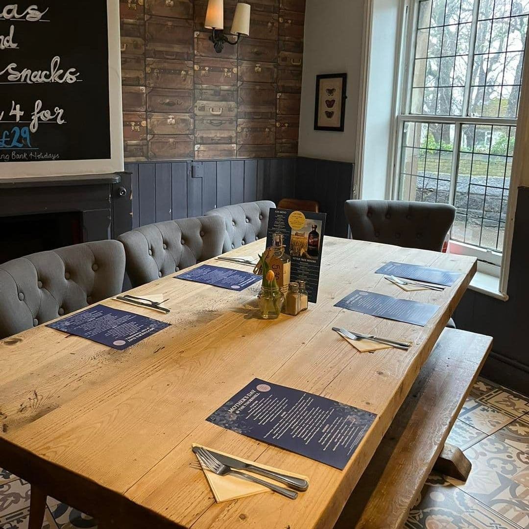 Interior of The Thornhill featuring a rustic dining setup with a long wooden table, set cutlery, menus, and condiments, with a cozy bench seating against a wall with decorative elements and a view of a window overlooking the exterior.
