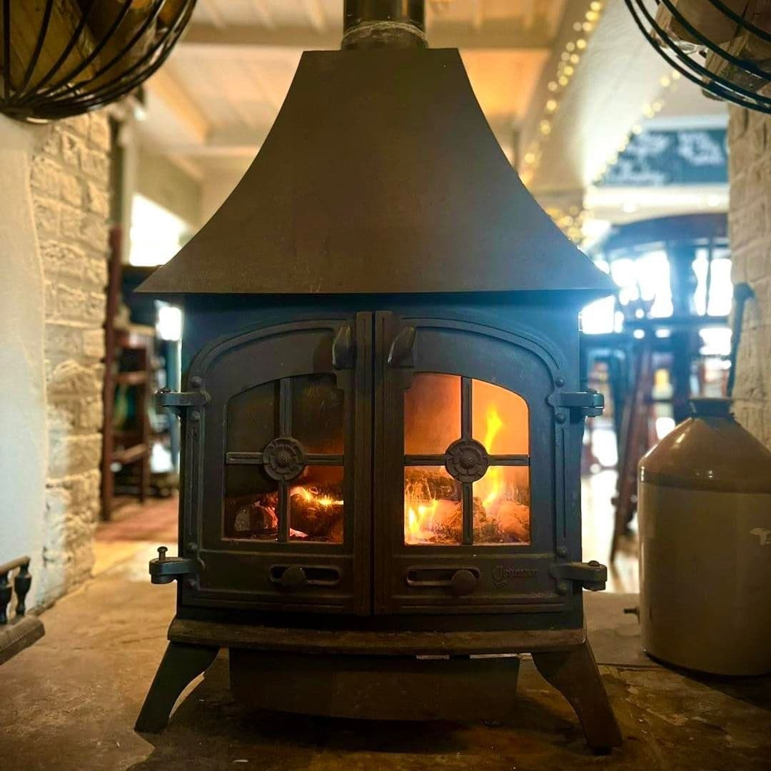 Cozy wood-burning stove with a warm fire at The Thornhill, set against a stone wall with a glimpse of the interior ambiance in the background.