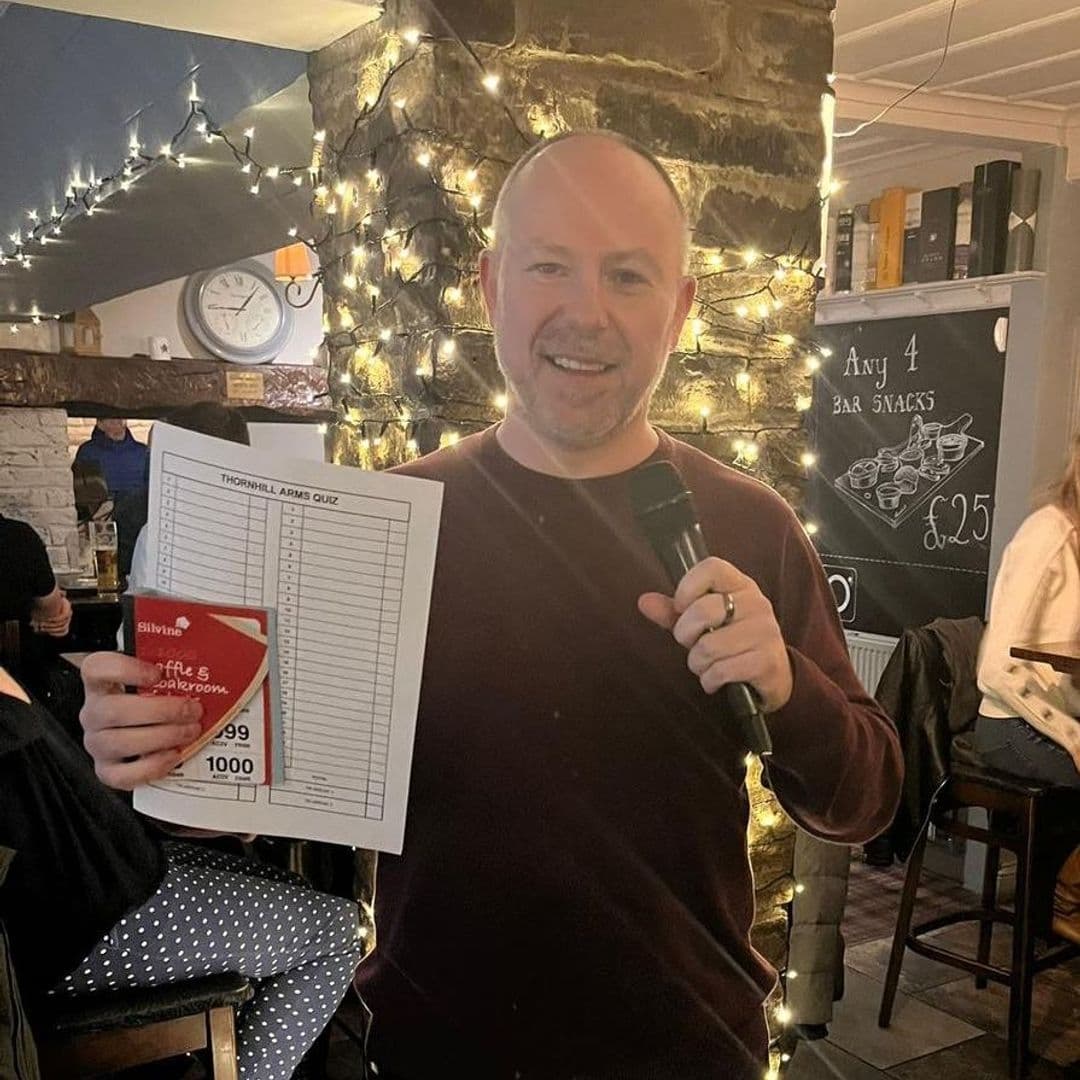 Man holding a quiz sheet and microphone at The Thornhill, with fairy lights and a chalkboard menu in the background.