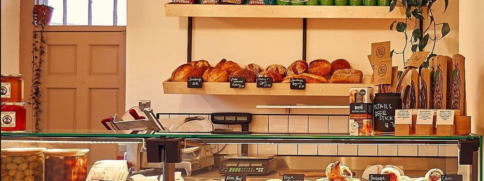 Artisanal bread display at Bottle Chop, featuring a variety of freshly baked loaves on wooden shelves, jars of honey, and gourmet snacks, with a deli slicer and cash register in the foreground.
