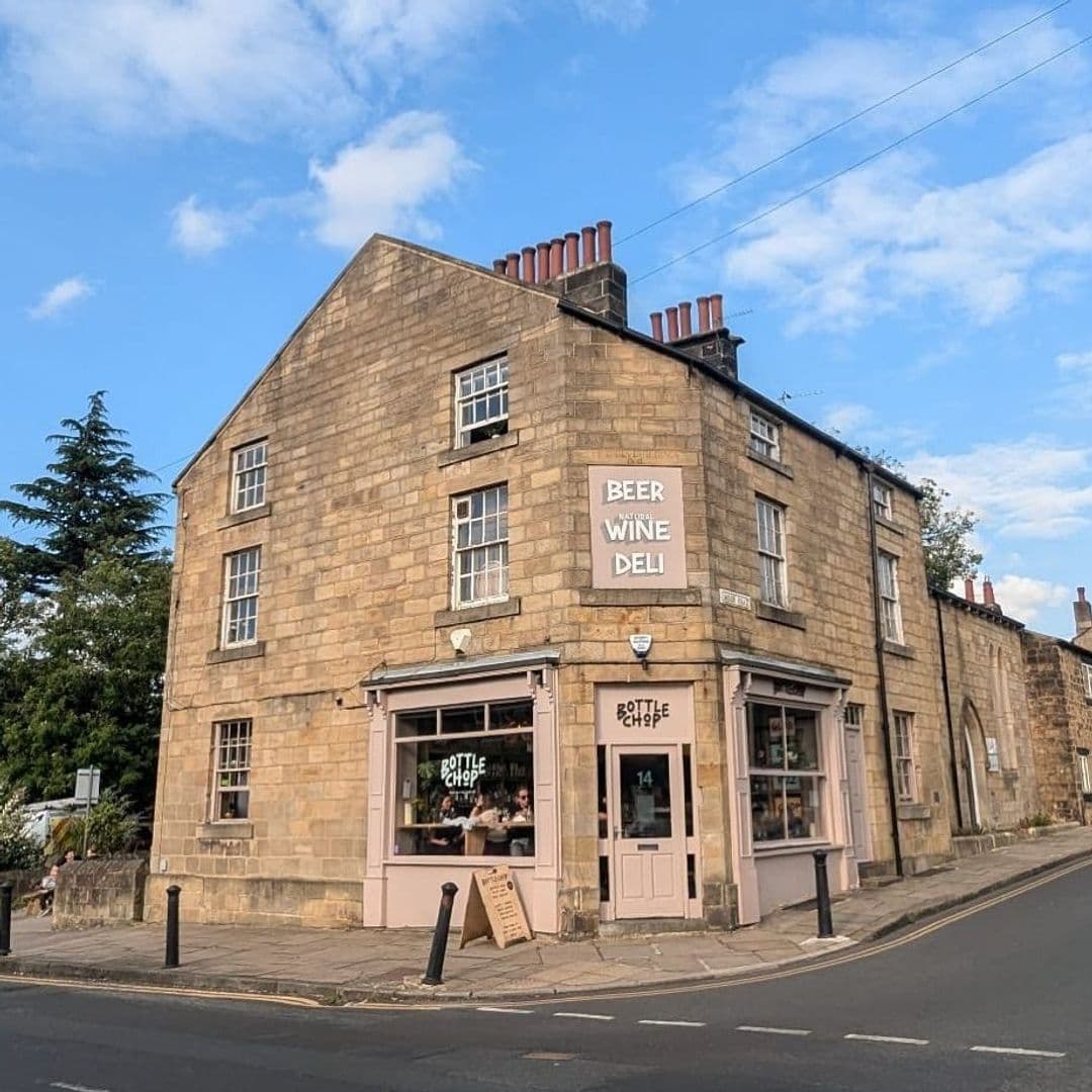 Alt text: "Exterior view of Bottle Chop, a corner building with a beige stone facade, featuring a beer, wine, and deli shop with large windows and a signboard outside, under a clear blue sky."