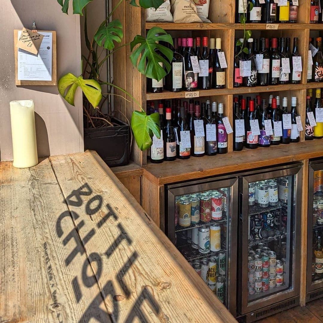 Interior view of Bottle Chop featuring a rustic wooden counter with the venue's name, a variety of wines displayed on shelves, and refrigerators stocked with canned beverages.