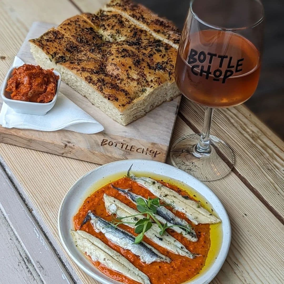 Alt text: "Gourmet appetizers at Bottle Chop featuring focaccia bread with za'atar seasoning, a side of red pepper spread, marinated white anchovies on a bed of tomato sauce, garnished with fresh herbs, accompanied by a glass of amber liquid with the Bottle Chop logo."
