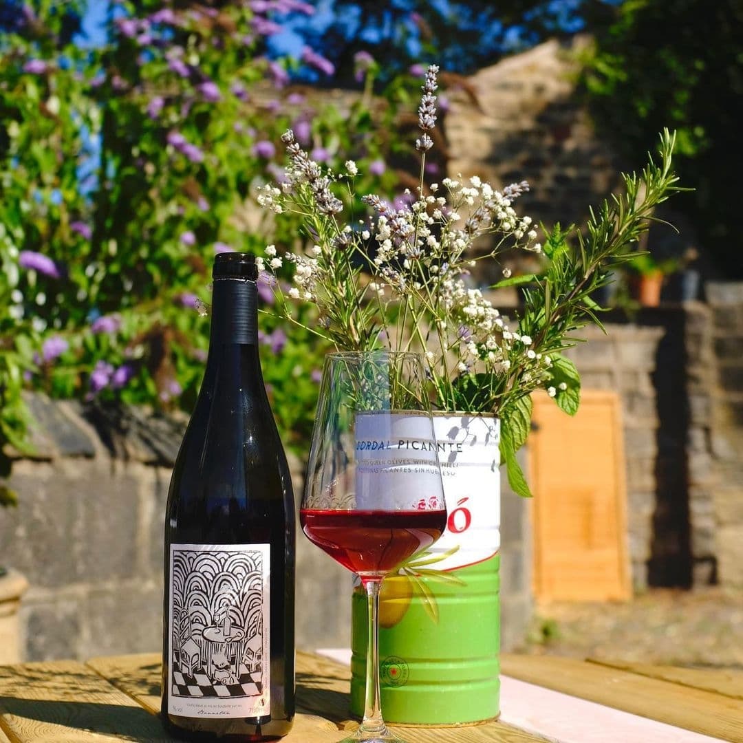 A bottle of red wine with a decorative label next to a wine glass half-filled with red wine, and a repurposed can vase with white flowers on a wooden table at Bottle Chop, with a blurred natural background suggesting an outdoor setting.