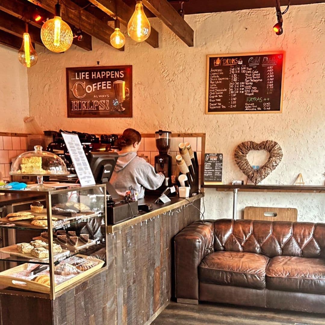 Interior of Cup 19 Coffee with barista at espresso machine, pastry display case, and menu boards on wall. Cozy seating area with leather sofa and decorative heart on wall.
