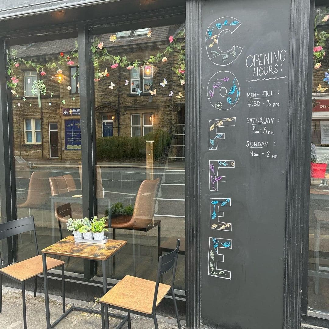 Exterior view of Cup 19 Coffee with outdoor seating, a blackboard displaying opening hours, and decorative lights and flowers above the window.