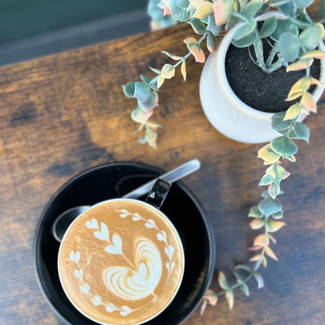 Artistic latte with heart and leaf pattern in a black cup at Cup 19 Coffee, accompanied by a potted succulent on a wooden table.