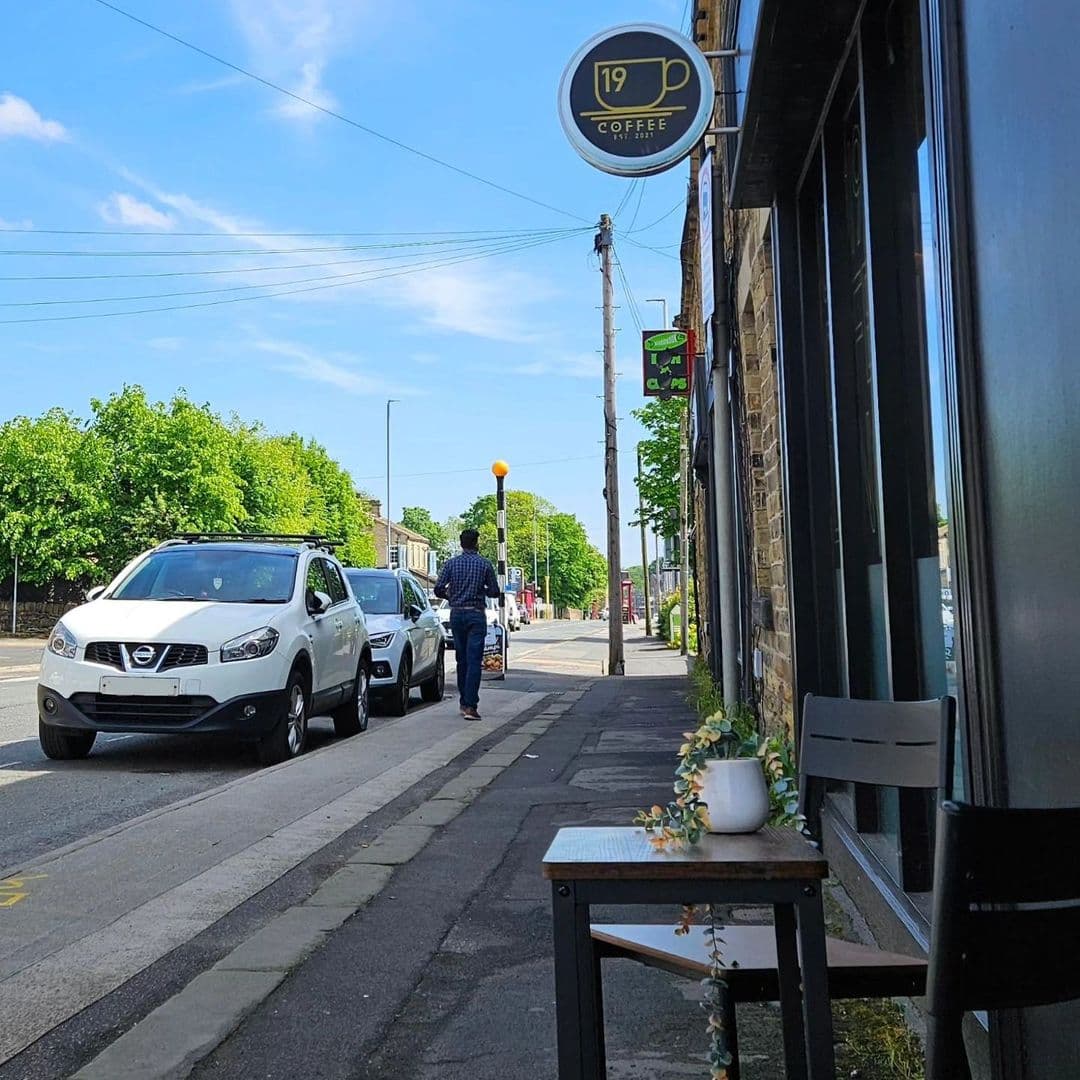 Street view outside Cup 19 Coffee with a clear blue sky, parked cars, a pedestrian, and the shop's round sign prominently displayed.