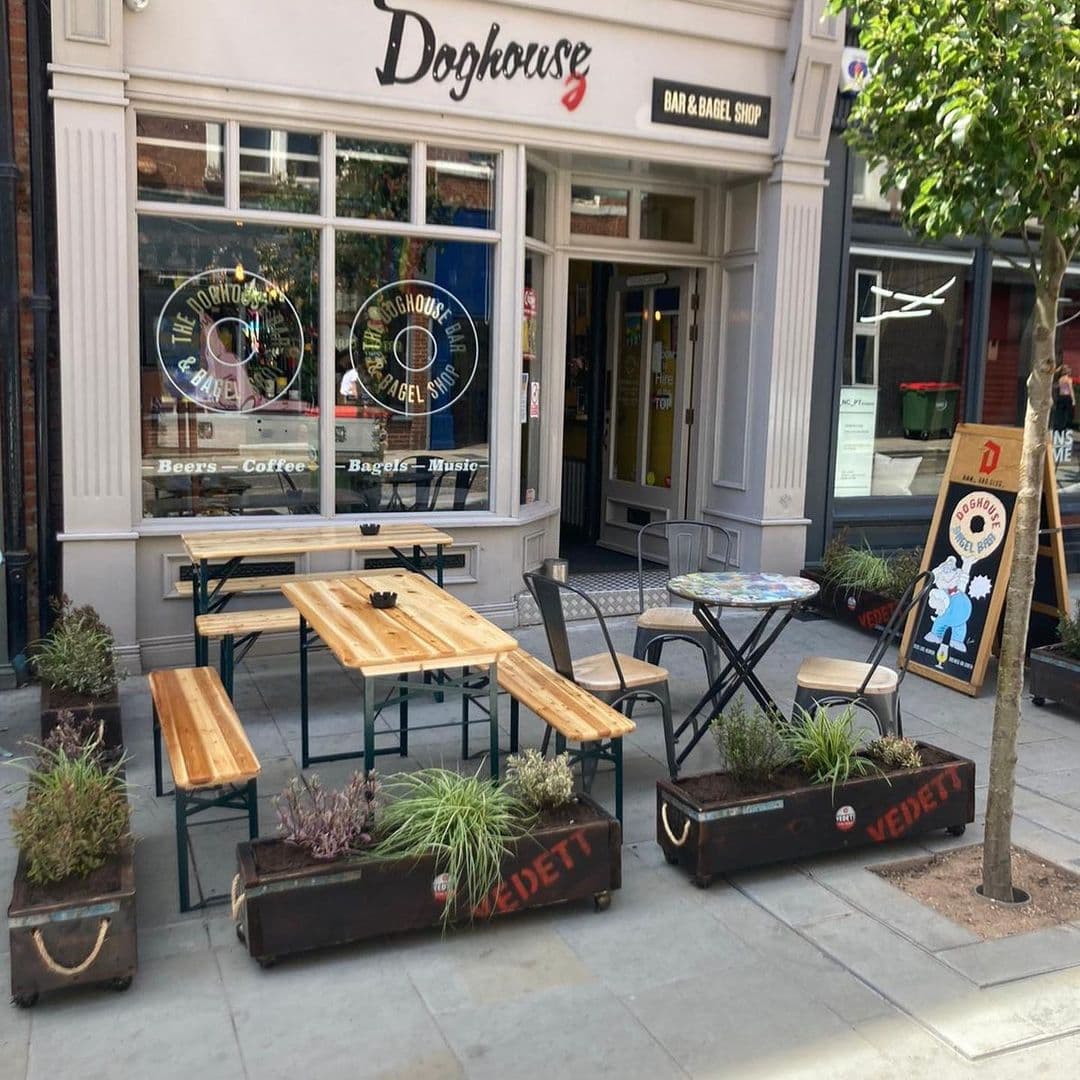 Exterior view of Doghouse Bar & Bagel Shop with outdoor seating, planters, and signage promoting beers, coffee, bagels, and music.