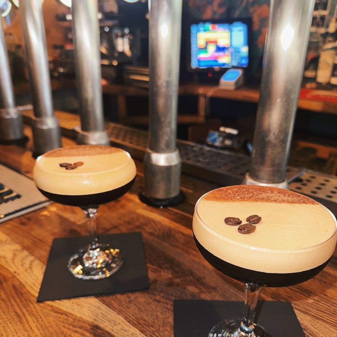 Two creamy espresso martinis on a bar counter at Doghouse Bar & Bagel Shop, with beer taps in the background and a slot machine screen visible. Each martini is garnished, one with a coffee bean and the other with three beans on its frothy top.