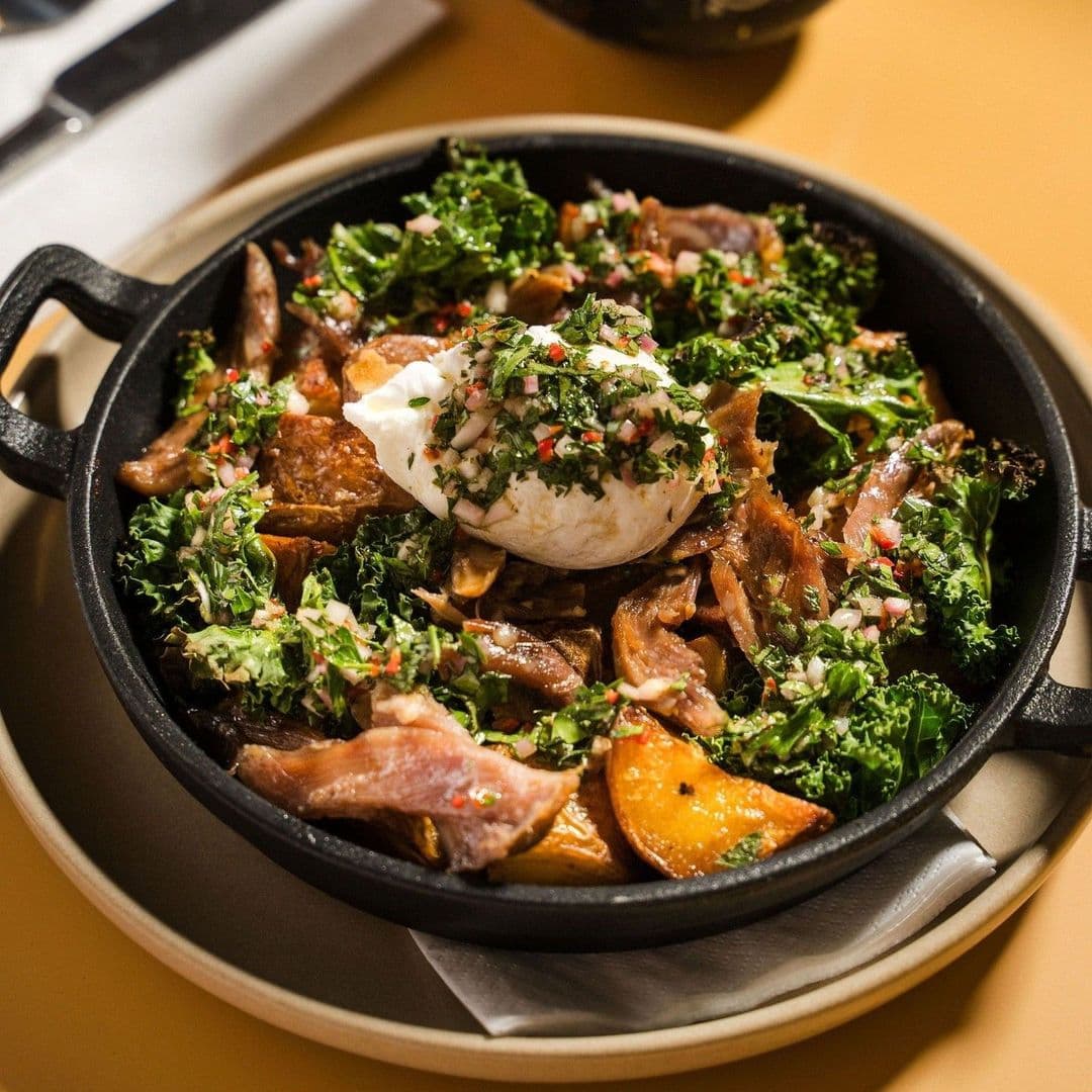 Savory dish served at Galleria featuring roasted meats, crispy kale, potatoes, and a poached egg garnished with fresh herbs in a cast-iron skillet.