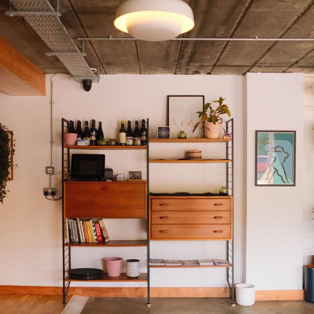 Interior view at Galleria featuring a modern shelving unit with assorted items including books, wine bottles, a vintage radio, and potted plants, with a framed artwork on the wall.