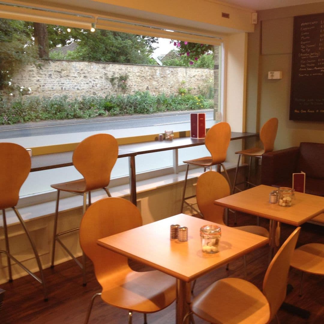 Interior of Maypole Coffee Shop & Deli with wooden tables and chairs, menu board on the wall, and a window view overlooking a street and stone wall.