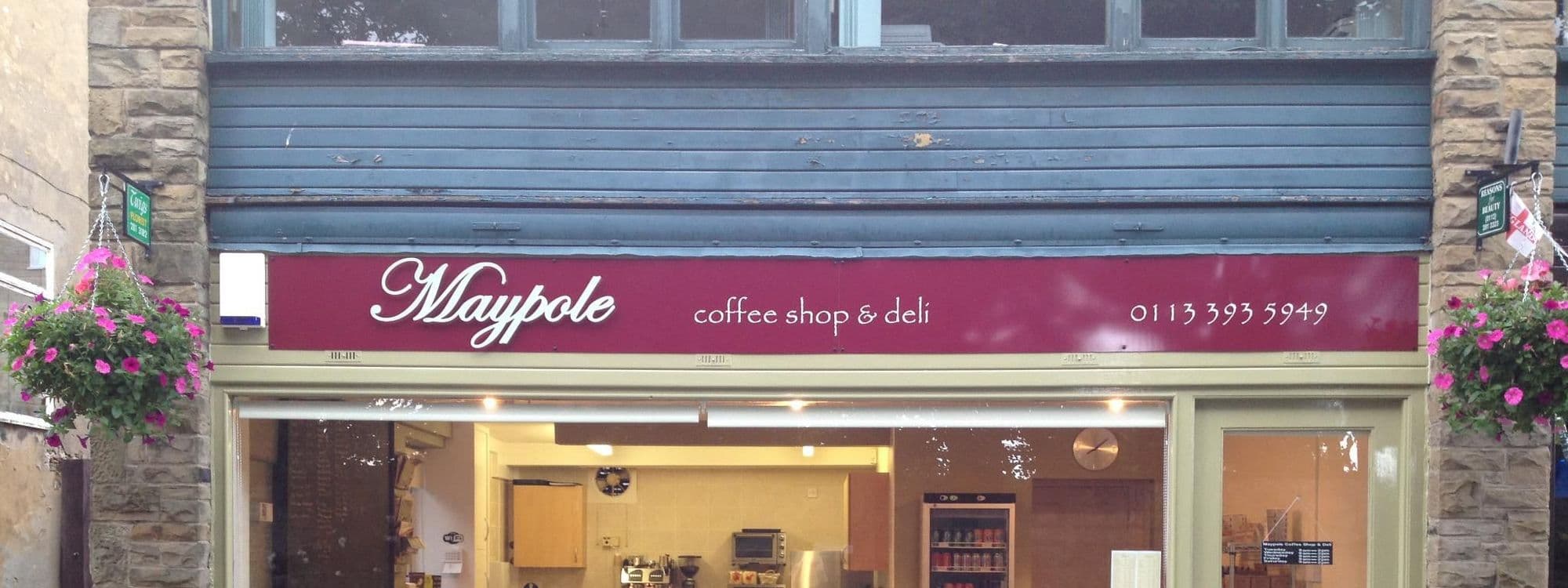 Exterior view of Maypole Coffee Shop & Deli with a burgundy awning, hanging flower baskets, and visible interior including counter and shelves.