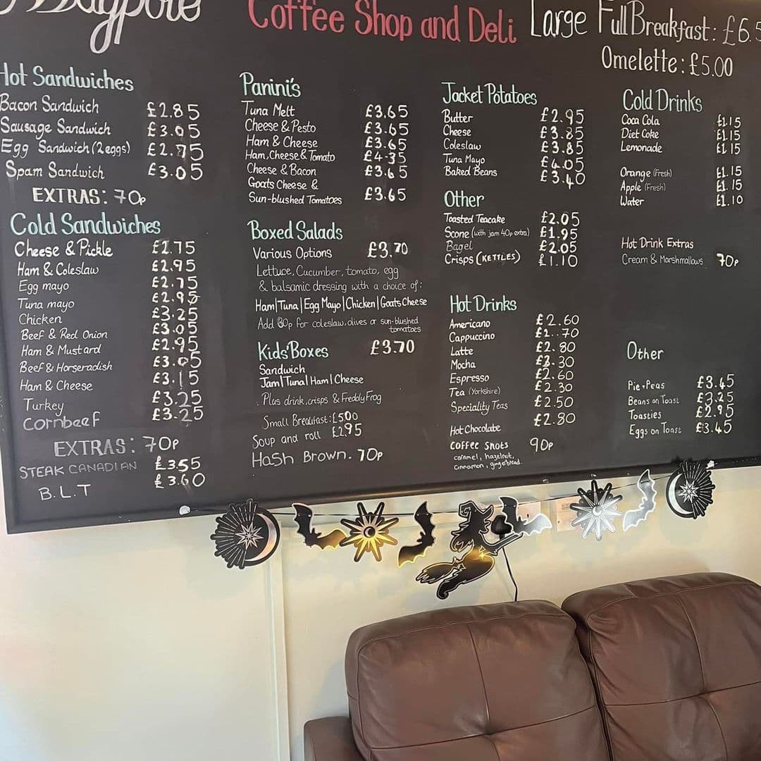 Interior of Maypole Coffee Shop & Deli featuring a menu board with various food and drink options such as hot sandwiches, paninis, salads, and hot drinks, with prices in British pounds. Comfortable brown seating is visible below the menu. Decorative elements include a blackboard with white chalk writing and wall-mounted metallic decorations with coffee and tea motifs.