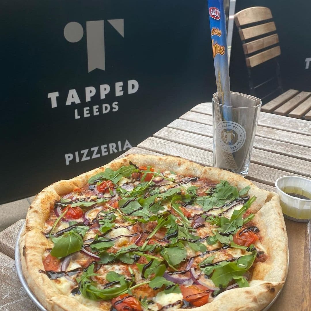 Freshly baked pizza with arugula and cherry tomatoes on a wooden table at Tapped Leeds Pizzeria, with a branded pint glass and outdoor seating in the background.