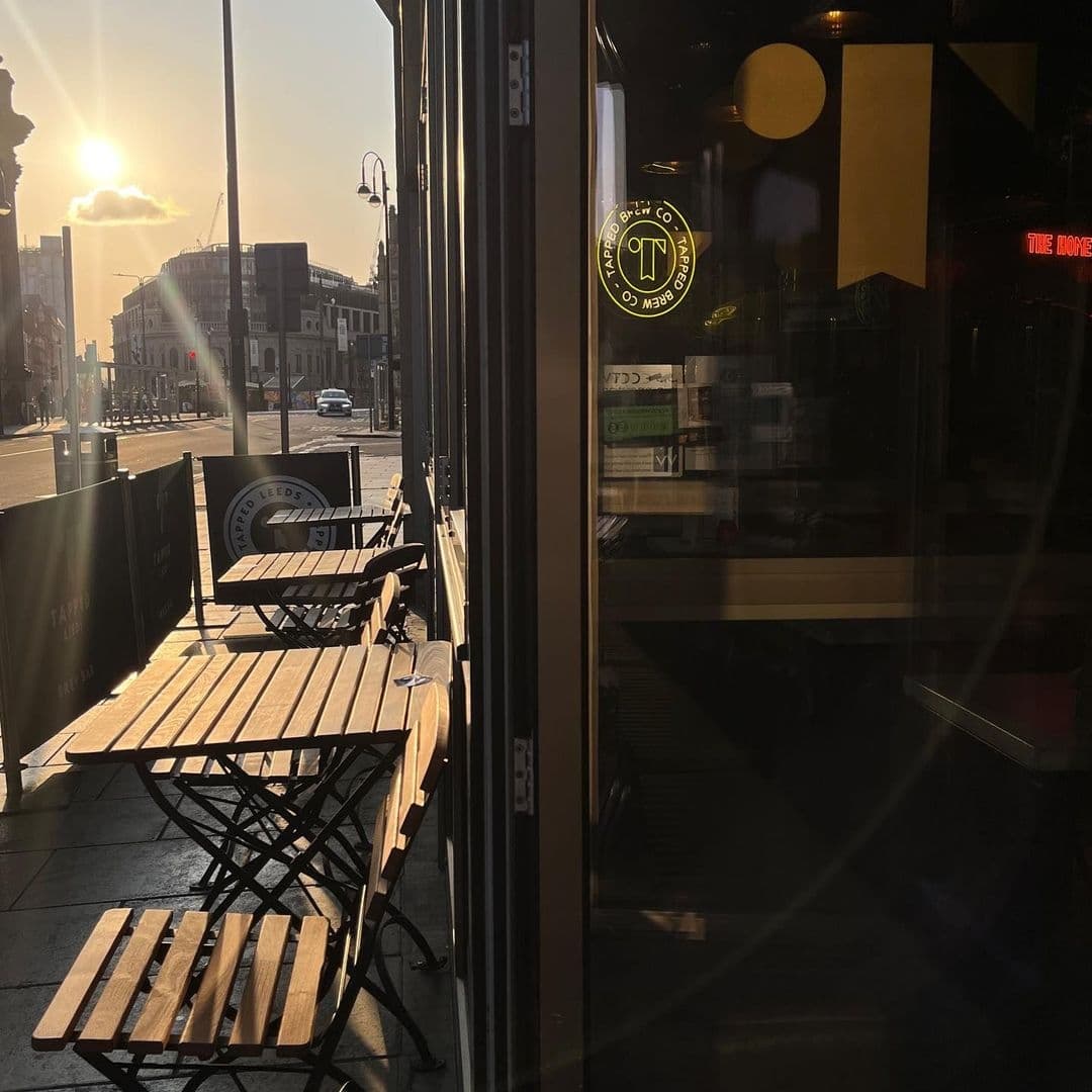 Sunlit outdoor seating at Tapped with empty wooden tables and chairs, city street in the background, and the establishment's logo visible on the glass door.
