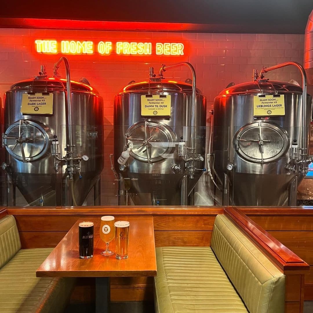 Interior of Tapped brewery with three large stainless steel beer fermentation tanks and a neon sign reading "THE HOME OF FRESH BEER" above. A wooden table with two glasses of beer sits in the foreground, flanked by green upholstered bench seating.