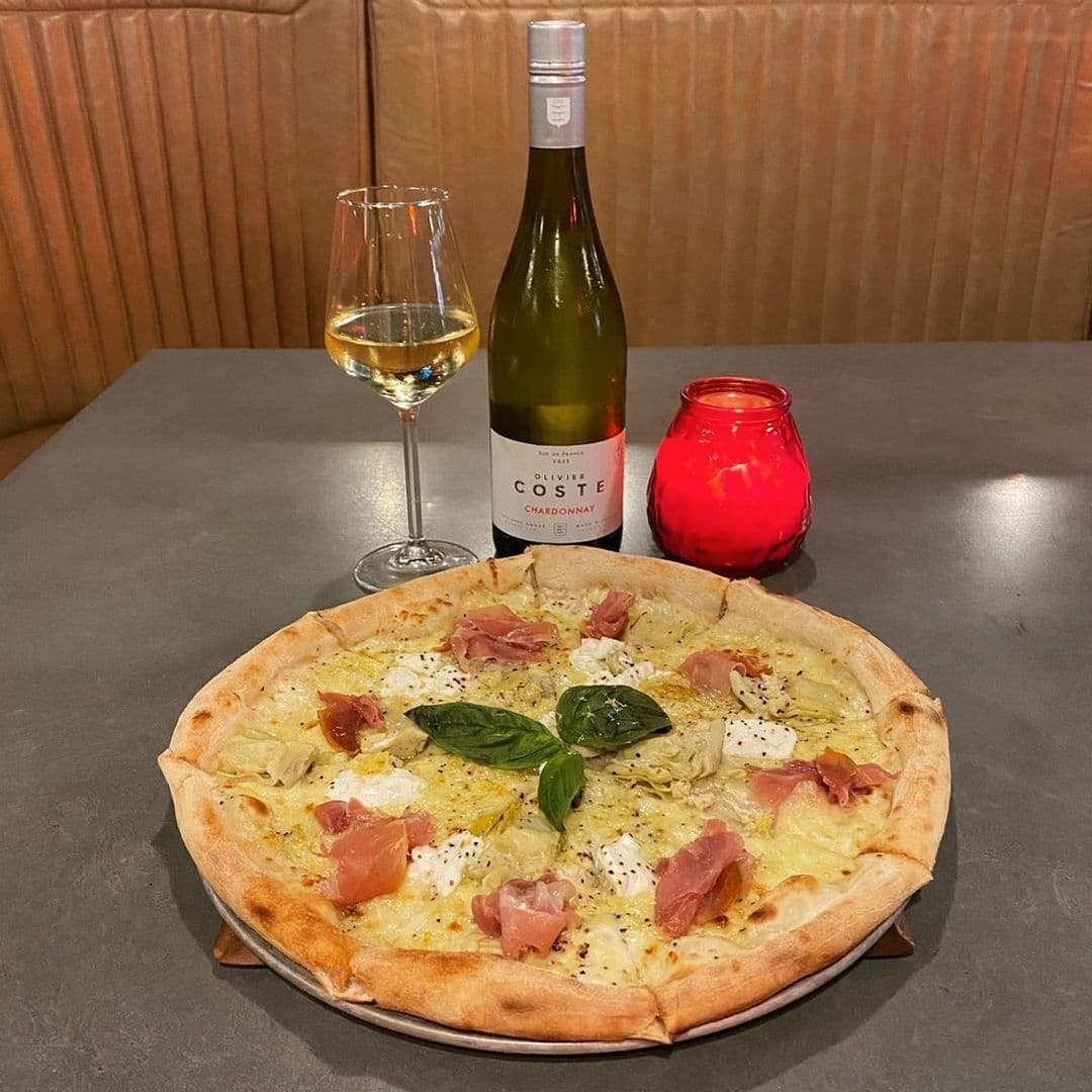 Gourmet pizza with prosciutto and basil on a table at Tapped, accompanied by a glass and bottle of Chardonnay wine and a red candle holder.