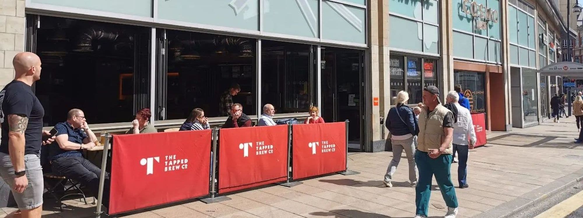 Outdoor seating at Tapped Brew Co with patrons enjoying drinks in the sun, surrounded by red barriers with the Tapped logo, on a lively city sidewalk.