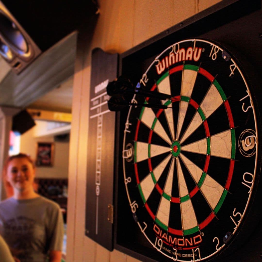Dartboard with darts in focus at The Eldon Pub, with a smiling patron in the background.