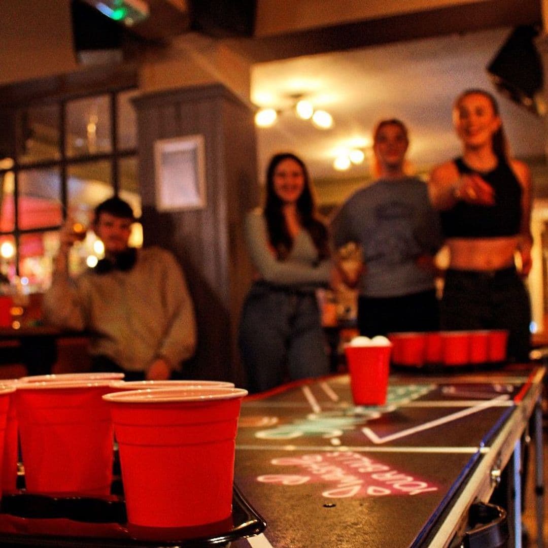 Patrons enjoying a game of beer pong at The Eldon Pub with red cups arranged on a table, in a cozy, warmly lit setting.