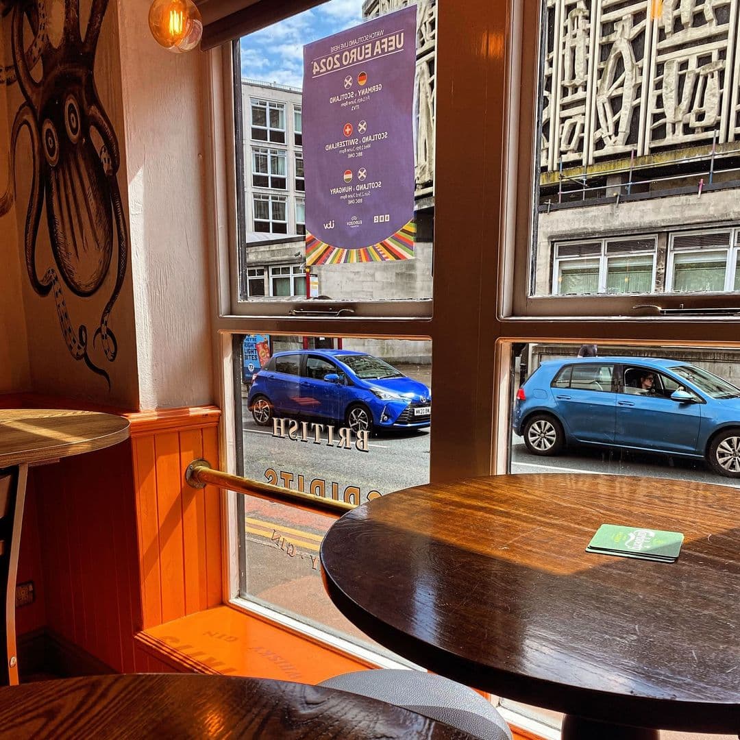 Interior view from The Eldon Pub with a round wooden table in the foreground, looking out onto a street through a window with "BITTER PILS" written on it, and a blue car visible outside.