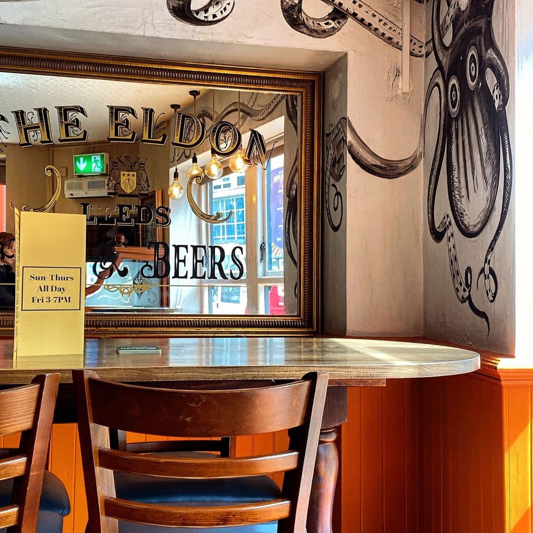 Interior of The Eldon Pub featuring a wooden table with chairs, a mirror with the pub's name and 'Leeds Beers' written on it, and an artistic wall mural of an octopus.