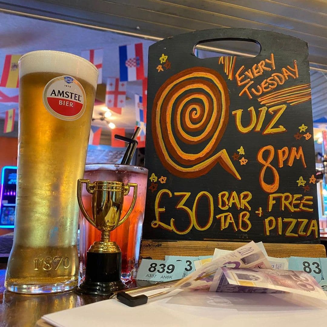 A pint of Amstel beer next to a cocktail and a small trophy, with a chalkboard advertising a quiz night at The Eldon Pub, featuring a £30 bar tab and free pizza. British currency notes and a pen are also visible on the table.