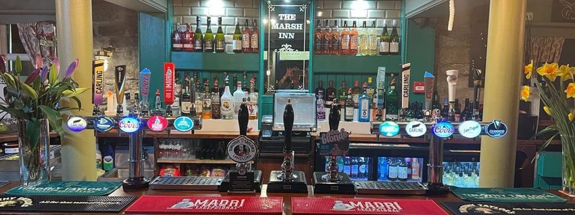 Bar counter at The Marsh Inn, Pudsey, displaying a variety of beer taps, spirits, and fresh flowers with the inn's name visible in the background.