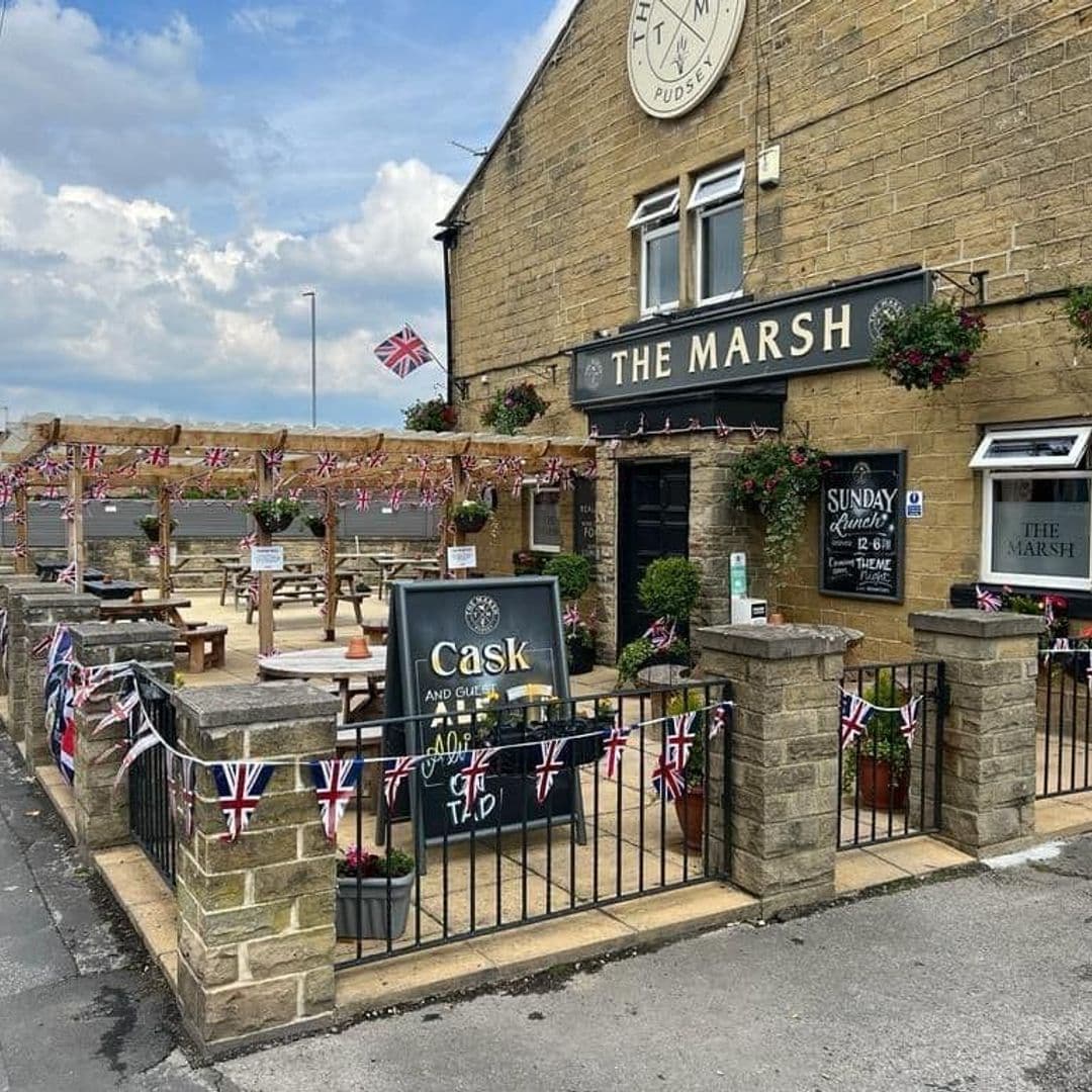 Exterior view of The Marsh pub in Pudsey with outdoor seating, British flags, and flower boxes under a clear sky.