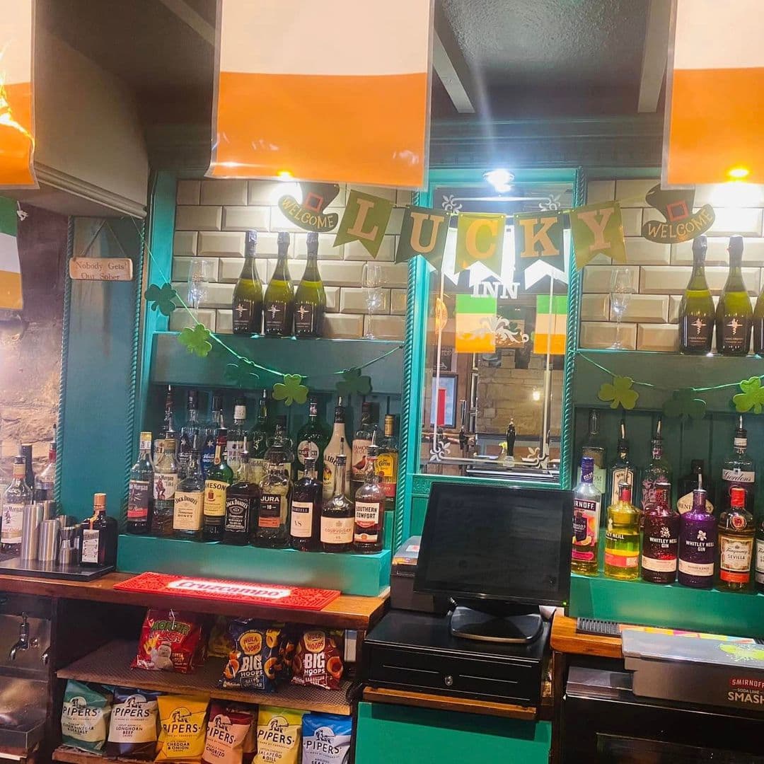 Interior of The Marsh – Pudsey showing a bar counter with a variety of spirits and snacks, decorated with green shamrocks and 'LUCKY INN' banners.