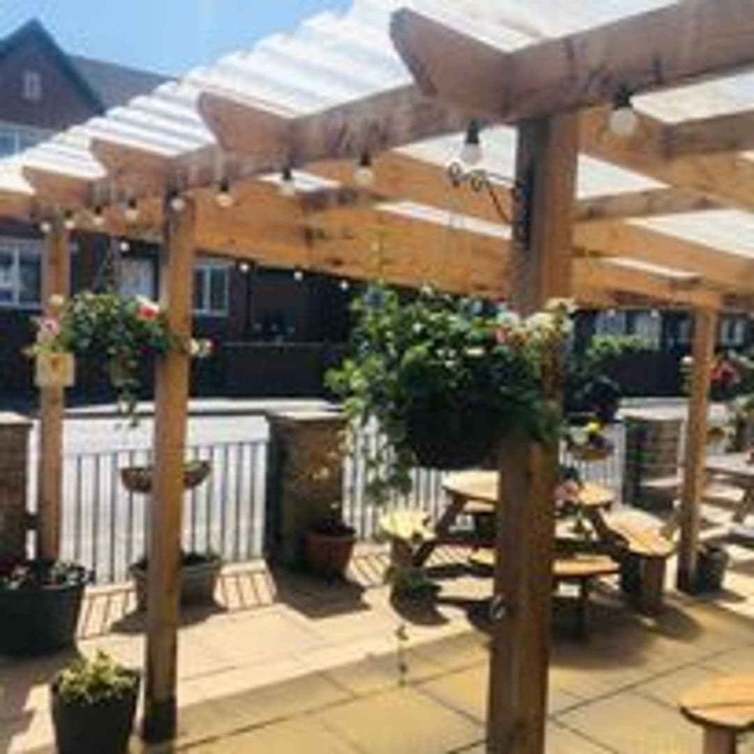 Outdoor seating area with wooden pergola and picnic benches at The Marsh – Pudsey, under a clear blue sky.