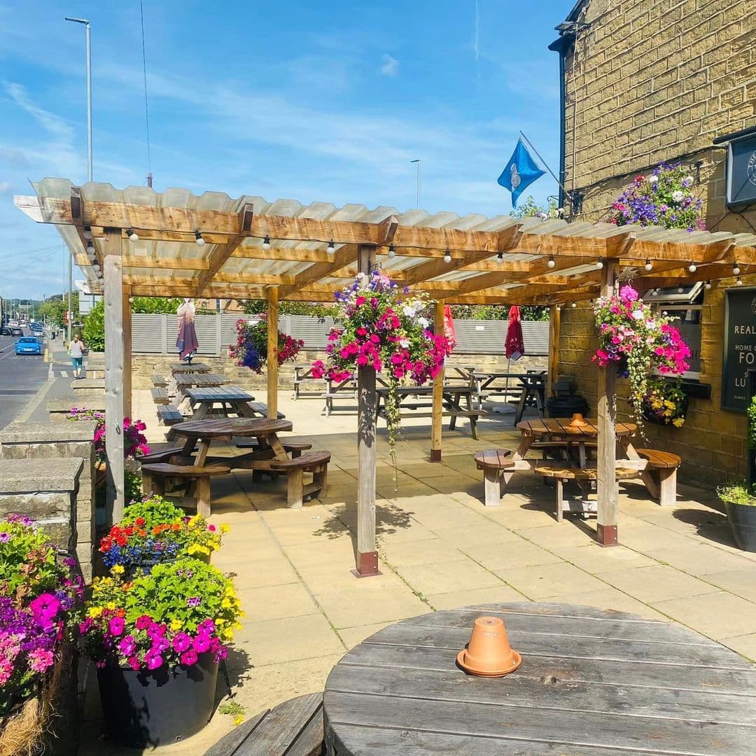 Outdoor seating area at The Marsh – Pudsey with wooden picnic benches, a pergola adorned with vibrant hanging flower baskets, and a clear blue sky.
