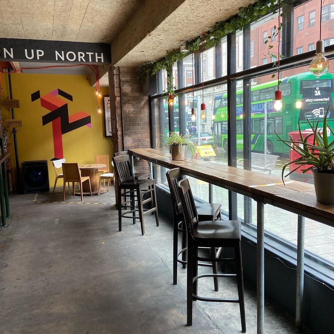 Interior view of The Old Red Bus Station, featuring a cozy seating area with high stools along a wooden bar table, a vibrant yellow wall with abstract art, and large windows overlooking green buses outside.