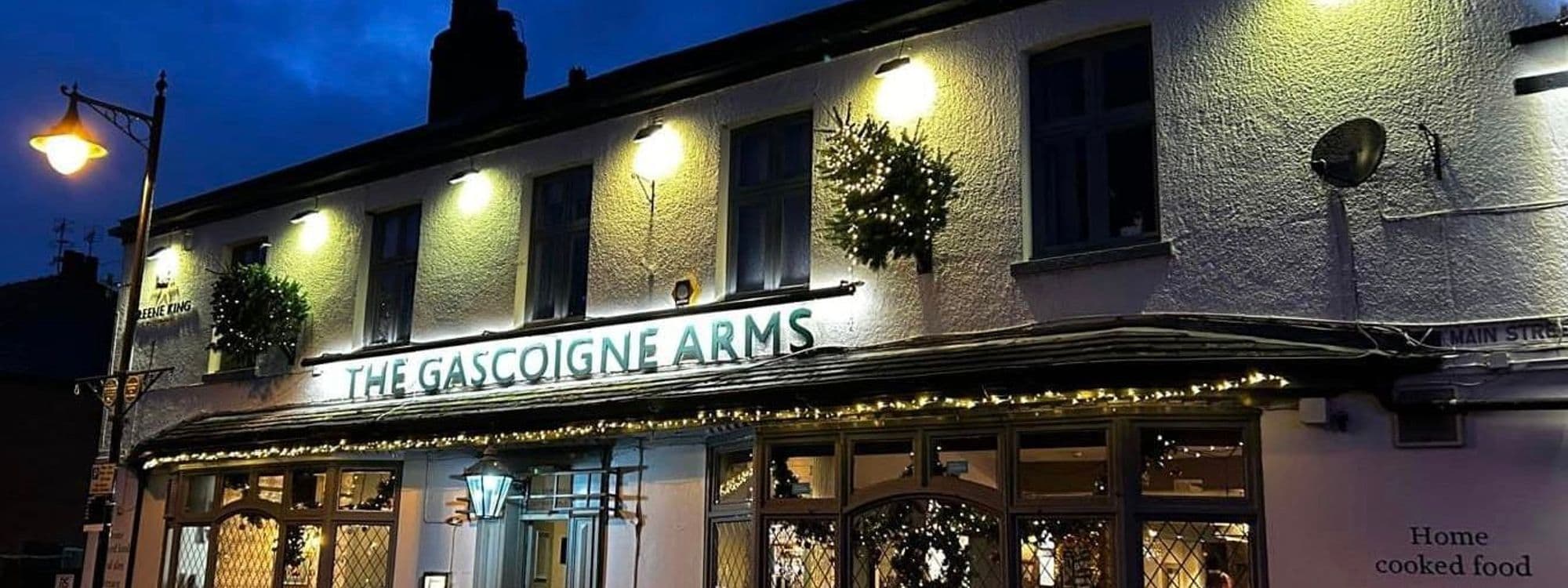 Exterior view of the Gascoigne Arms at dusk, showcasing its illuminated sign and festive lights around the windows and awning.