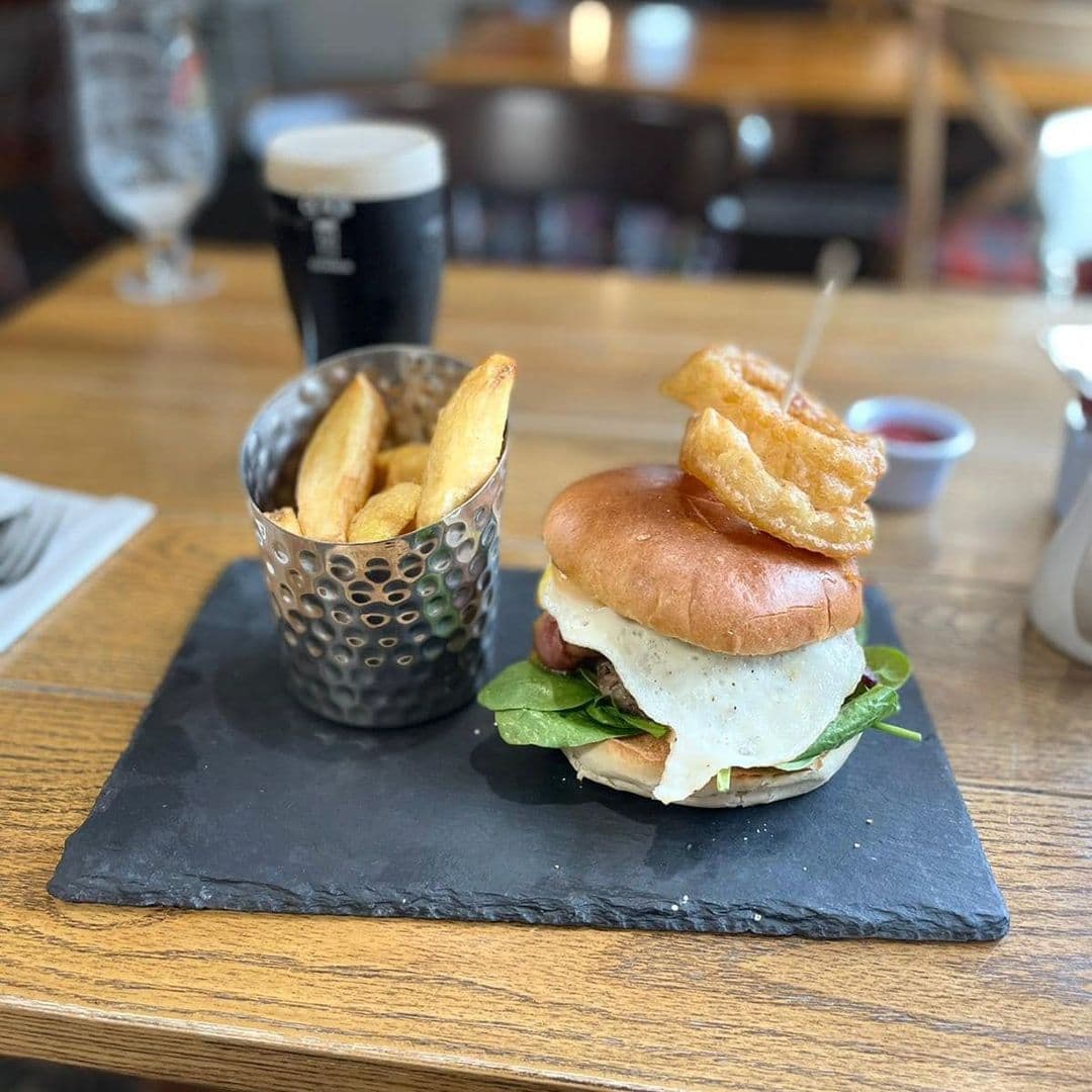 Gourmet burger with melted cheese and fresh greens on a brioche bun, served with thick-cut fries in a metal cup and a pint of dark beer at Gascoigne Arms.