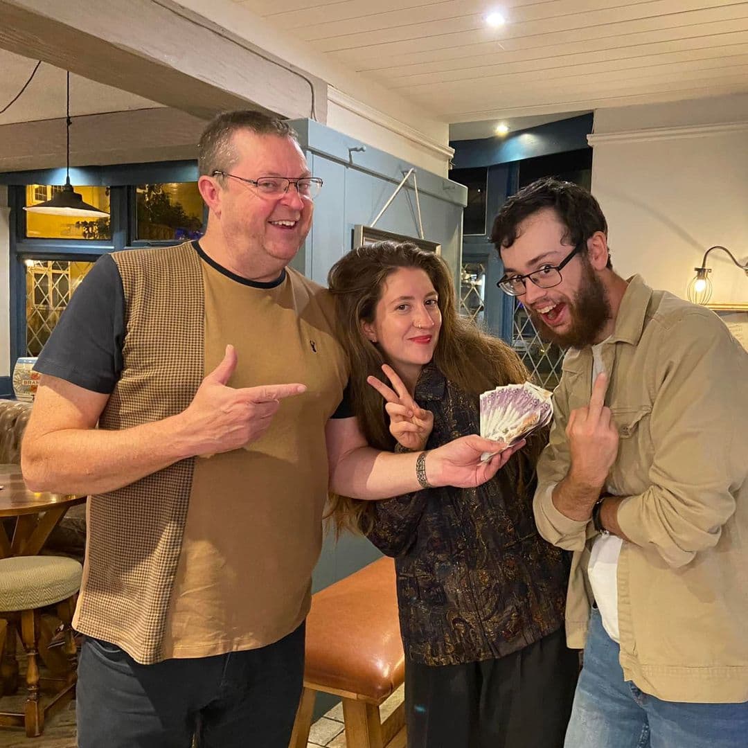 Three happy people posing with cash at the Gascoigne Arms, one man pointing at the woman in the center who is holding money, with another man giving a thumbs-up sign.