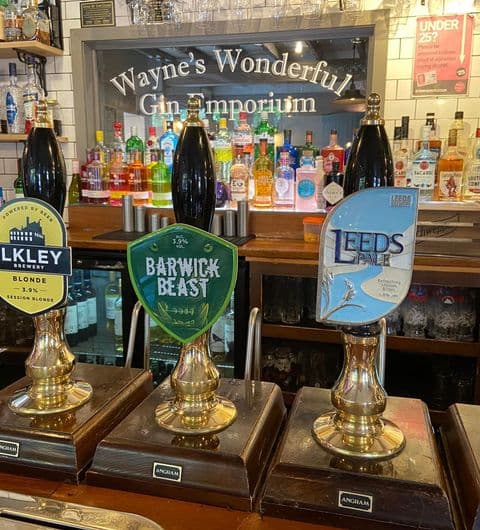 Traditional beer taps in the Gascoigne Arms featuring local brews, with a backdrop of a variety of gin bottles and a sign reading "Wayne's Wonderful Gin Emporium."