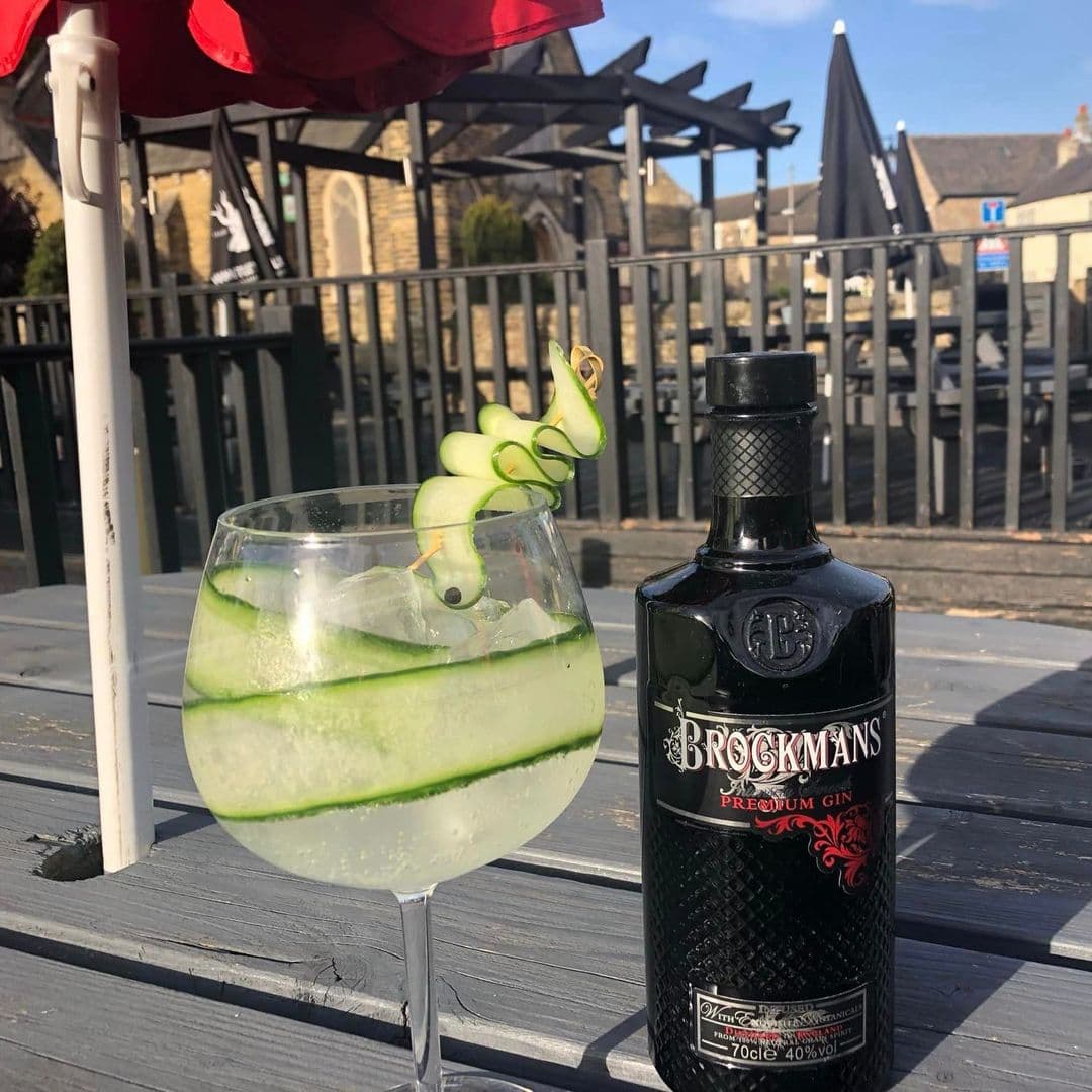 Outdoor dining at Gascoigne Arms featuring a gin cocktail with cucumber garnish and a bottle of Brockmans Premium Gin on a wooden table, with patio umbrellas and fencing in the background.
